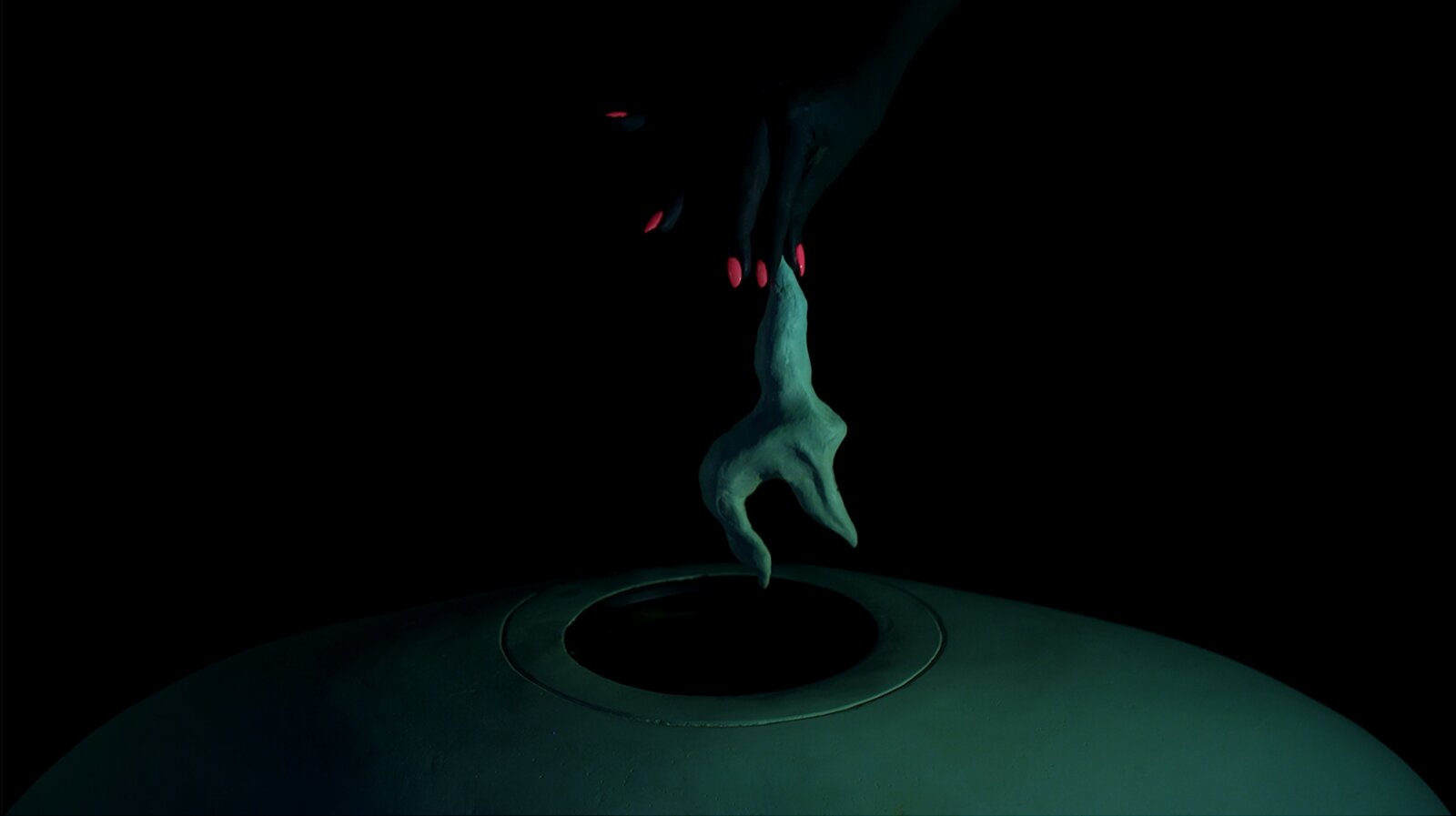 Still from Your Storm Our Dew by Cleo Fariselli: a dark hand with red nails holding a small green clay form above a round opening